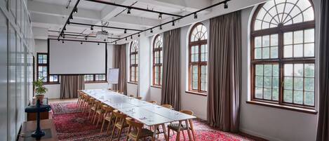 A conference room with a long table, chairs, a projector screen, and large windows with curtains.
