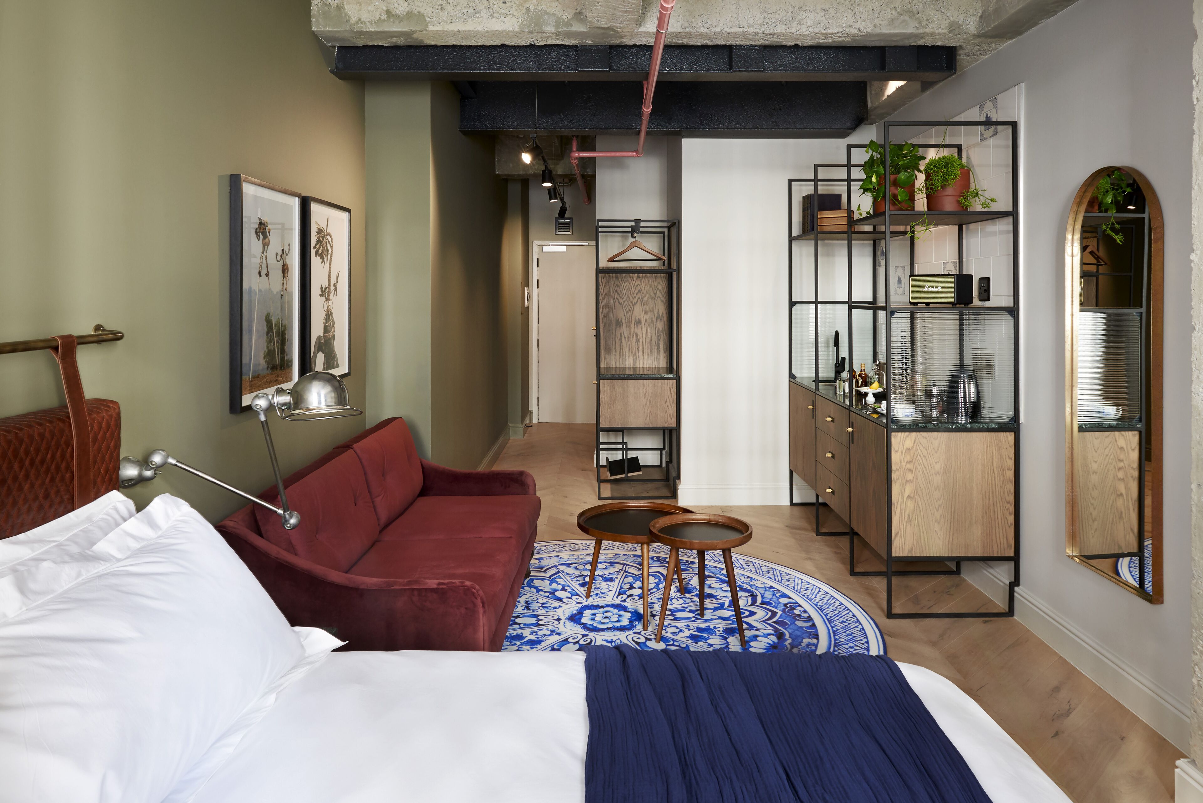 A modern hotel room with a bed, a red sofa, a small round table, a blue patterned rug, a wooden cabinet with shelves, and a mirror.