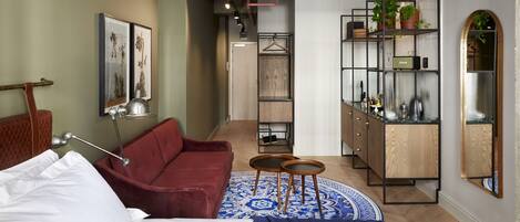 Quarto de hotel moderno com uma cama, um sofá vermelho, uma mesinha redonda, um tapete azul estampado, um armário de madeira com prateleiras e um espelho.