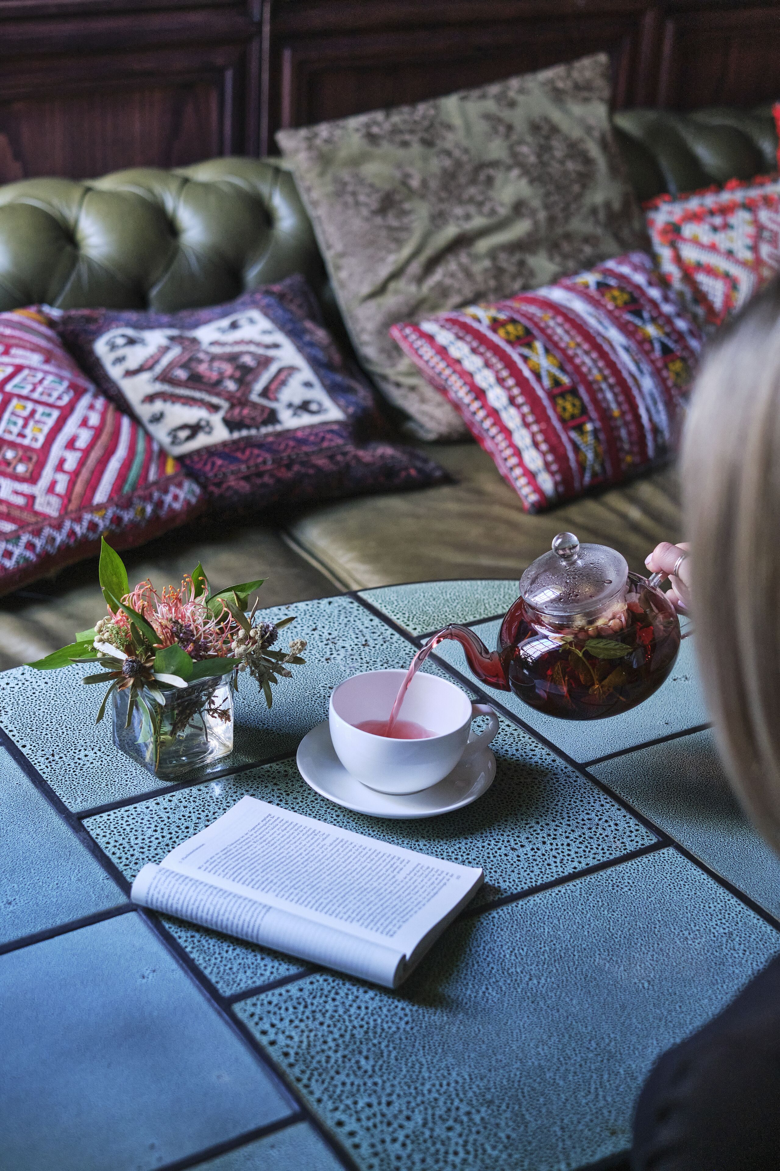 A cozy seating area with a green cushioned sofa, patterned pillows, a glass teapot, a cup, and a book on a tiled table.