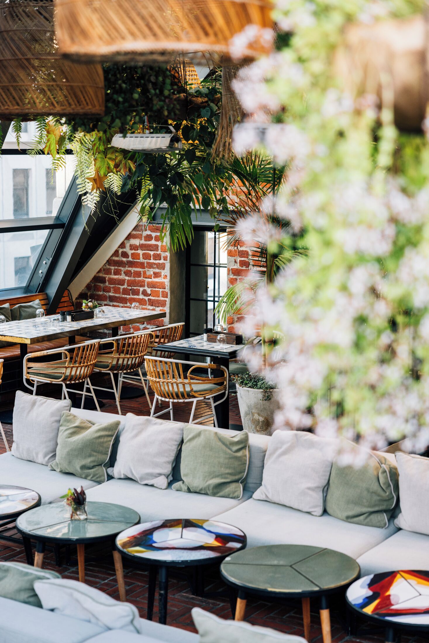 An outdoor seating area with wicker chairs and a cushioned bench, surrounded by potted plants and a brick wall.