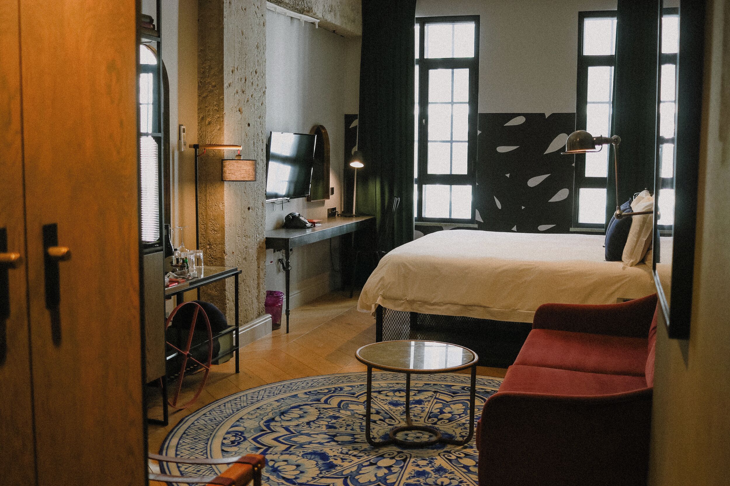 A hotel room with a bed, a desk with a TV, a chair, a small table, and a patterned rug.