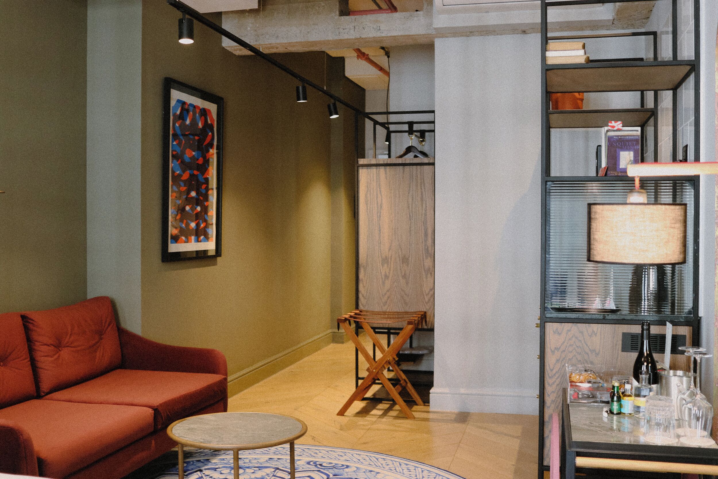 Un salon moderne avec un canapé rouge, une petite table ronde et une chaise en bois. On y trouve une bibliothèque remplie de livres et d’objets décoratifs, ainsi qu’un tableau abstrait encadré accroché au mur.