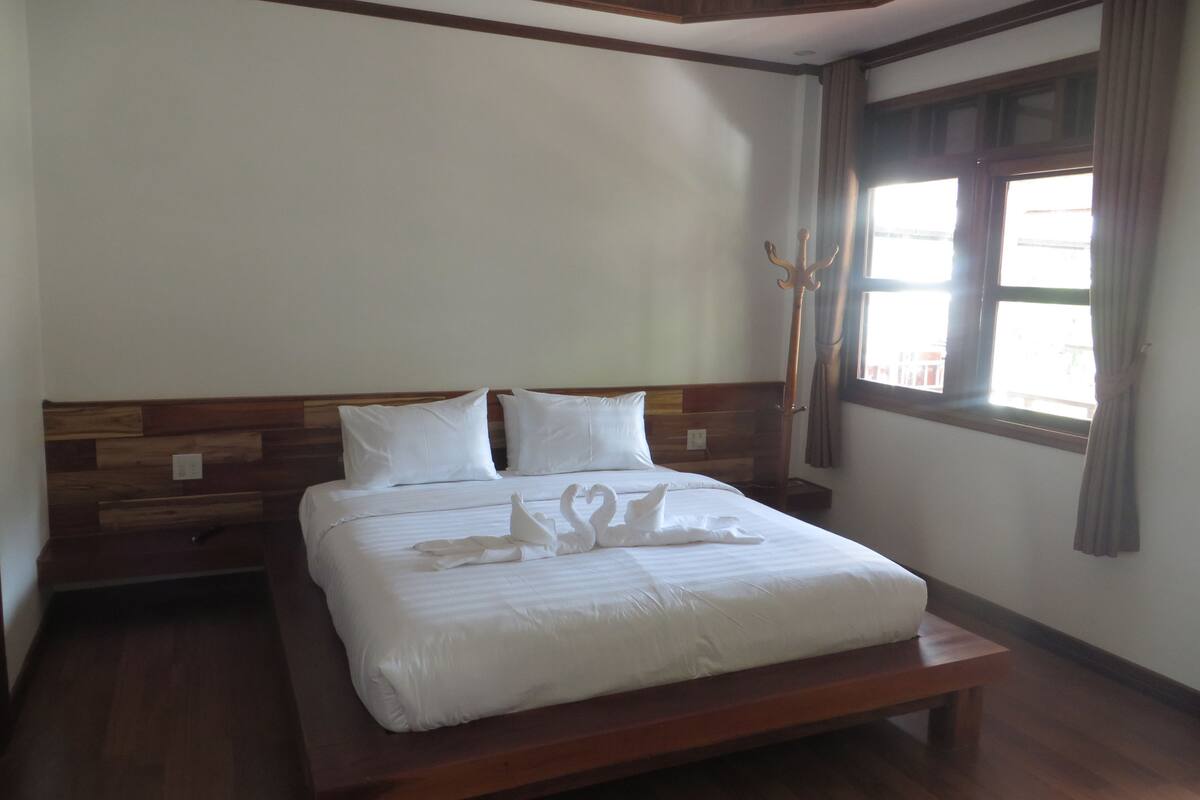 A neatly made bed with a wooden headboard, a wooden bench at the foot of the bed, and a window with curtains.