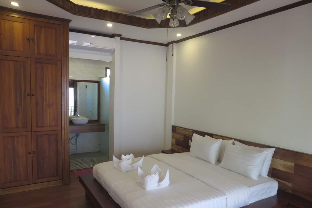 A bedroom with a wooden wardrobe, a bed with white linens and towels, and a bathroom visible through an open door.