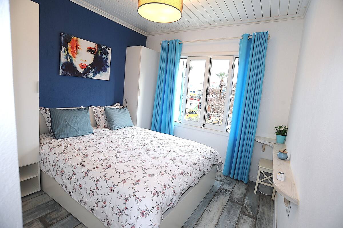 A bedroom with a bed, a nightstand, a window with curtains, and a painting on the wall.