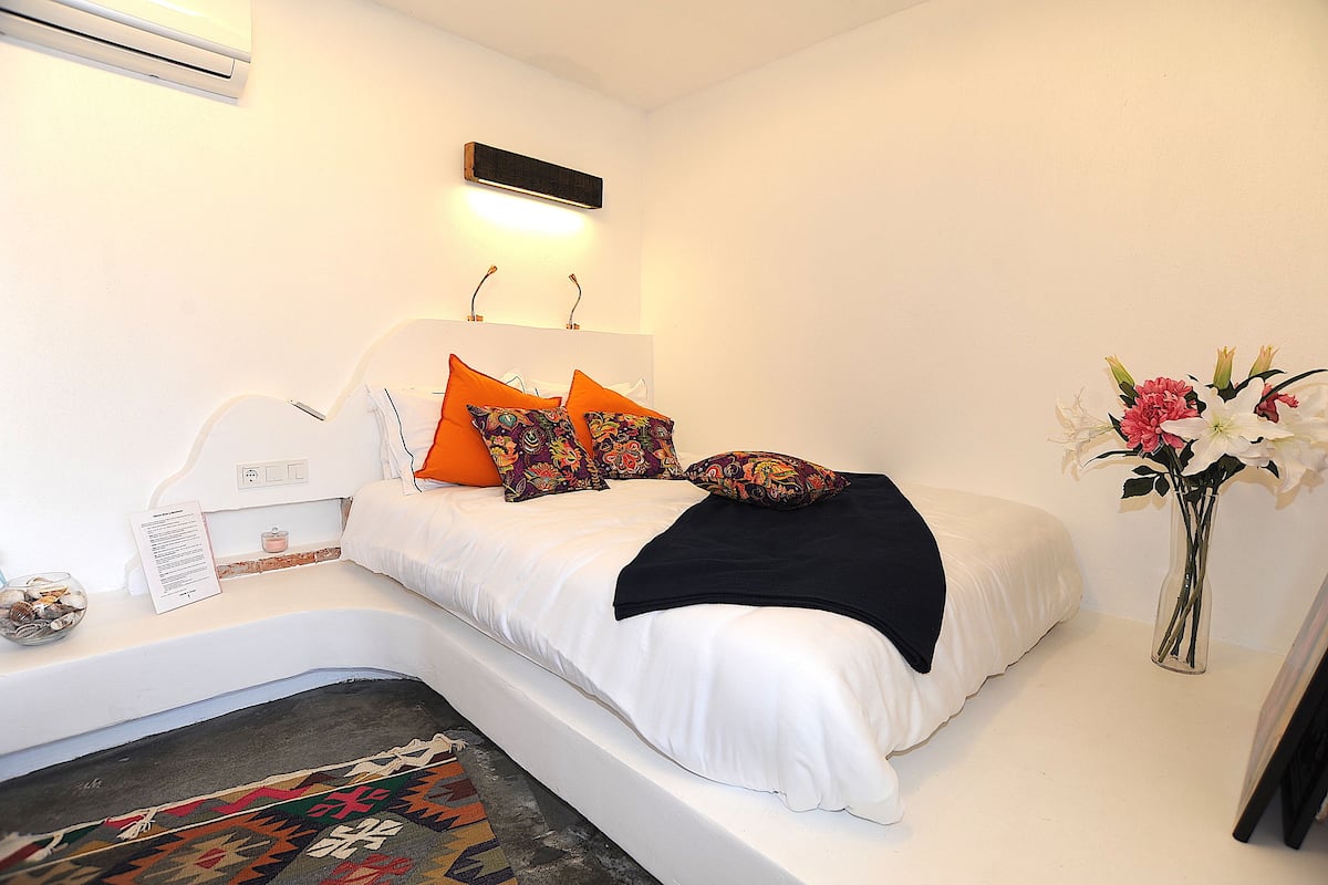 A neatly made bed with white bedding and colorful pillows, a small bedside table with a lamp, and a vase with flowers on the floor beside the bed.
