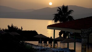 Vista del atardecer sobre un cuerpo de agua tranquilo, con una zona de playa que incluye sombrillas y asientos.