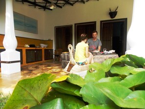 A covered outdoor area with a stone bench, a dining table, and a large plant in the foreground.