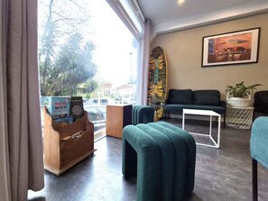 A room with a sofa, ottoman, and a surfboard leaning against the wall. - Balea Hostel (San Sebastián)