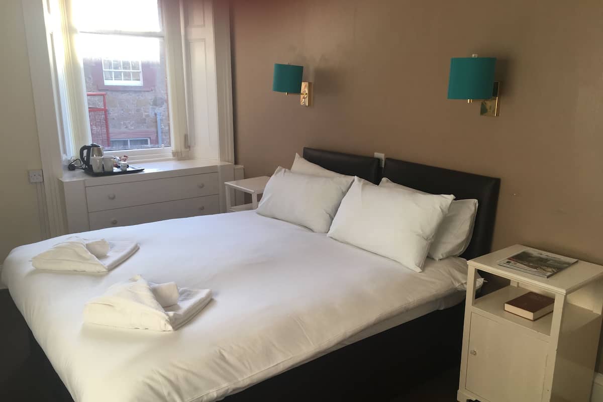 A neatly made bed with white linens and pillows, a bedside table with a book, and a window with a view of a building outside.