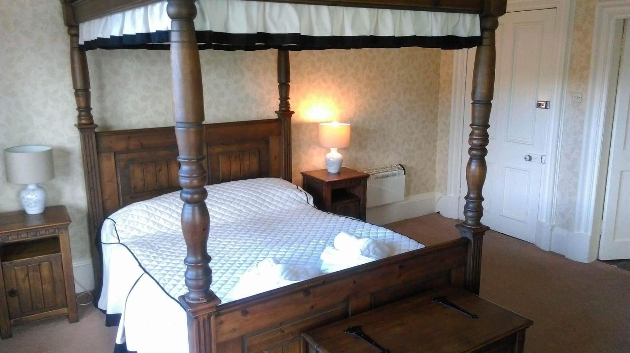 A four-poster bed with a quilted bedspread, a bedside table with a lamp, and a nightstand with a lamp.