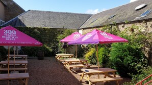 An outdoor seating area with picnic tables and umbrellas, surrounded by greenery and stone buildings. - George and Abbotsford Hotel (Melrose)