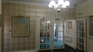 Vestíbulo de hotel con una araña, un mapa en la pared y un árbol de Navidad visible a través de las puertas de cristal.