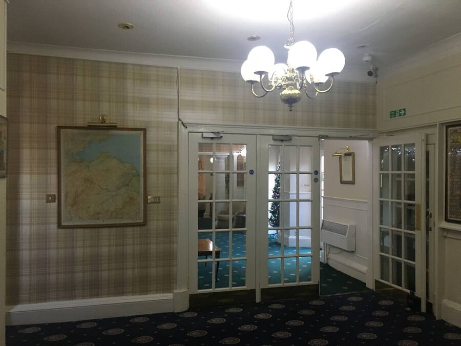 Vestíbulo de hotel con una araña, un mapa en la pared y un árbol de Navidad visible a través de las puertas de cristal.