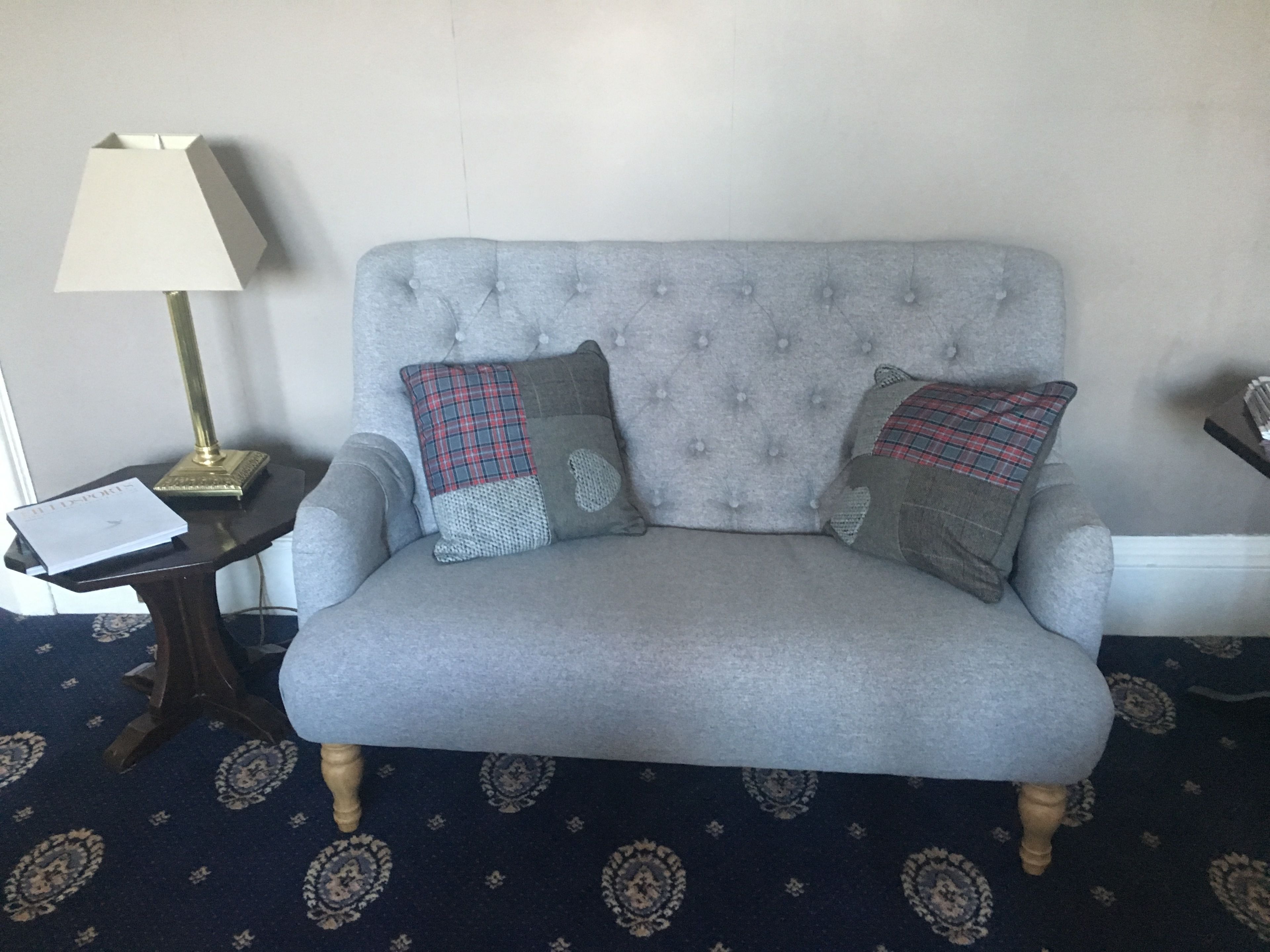 A grey upholstered sofa with tufted detailing and two decorative pillows, a small side table with a lamp, and a patterned carpet.