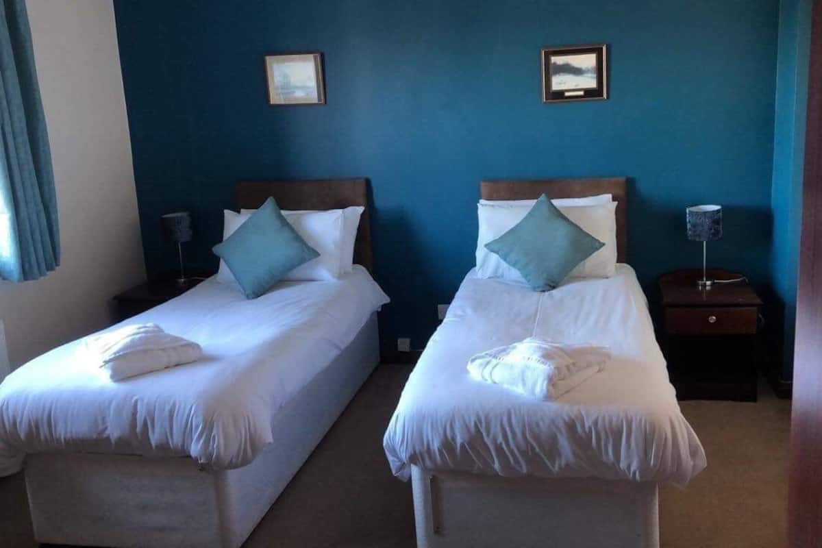 Two single beds with white linens and teal pillows, a nightstand with a lamp, and a blue wall with two framed pictures.