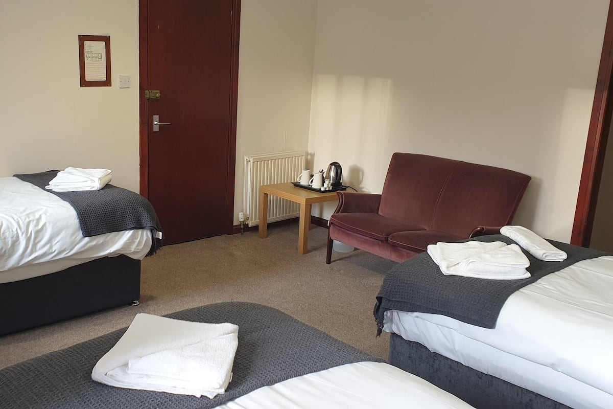 A hotel room with two beds, a red armchair, a small table with a kettle, and a framed picture on the wall.