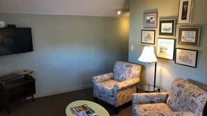 Ein Wohnzimmer mit Flachbildfernseher, zwei gemusterten Sesseln, einem kleinen runden Tisch mit einer Zeitschrift und einer Stehlampe.