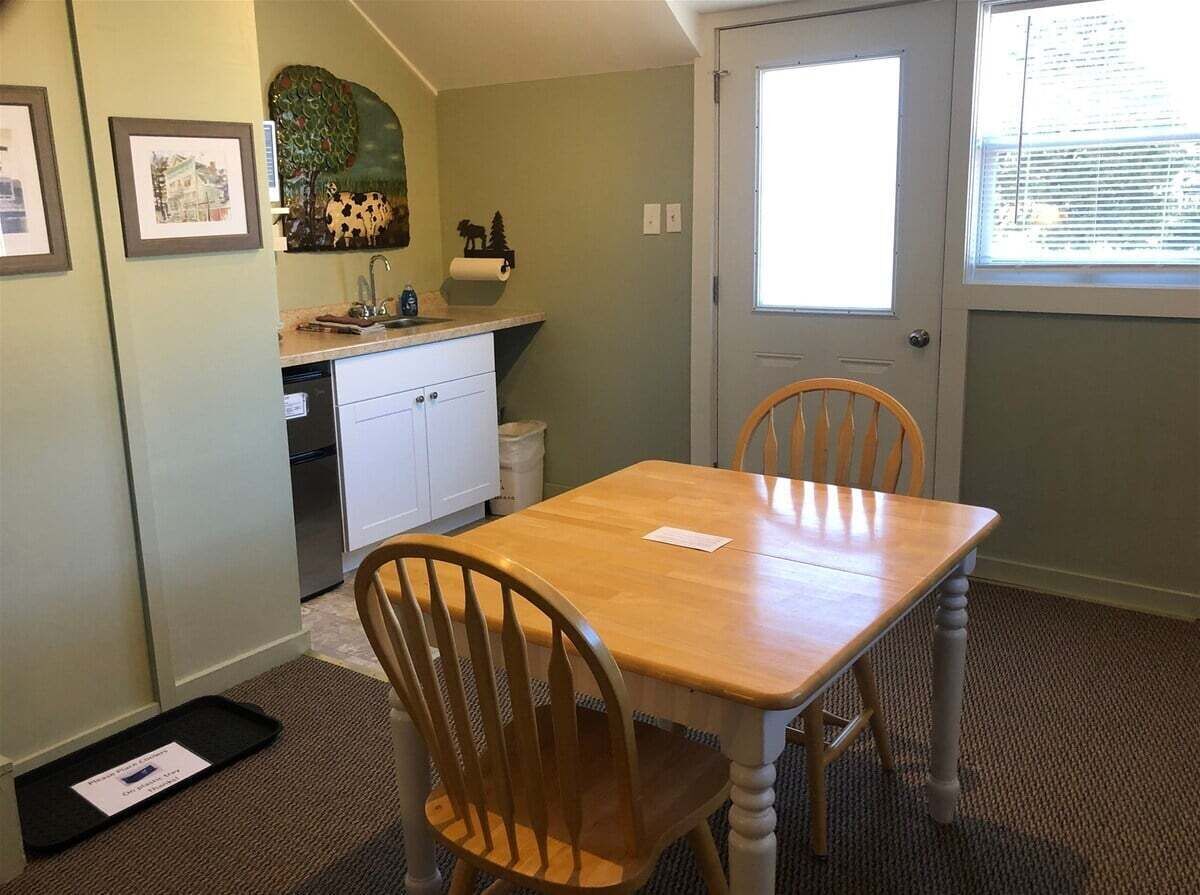 A dining area with a wooden table and chairs, a kitchen in the background, and a window with blinds.