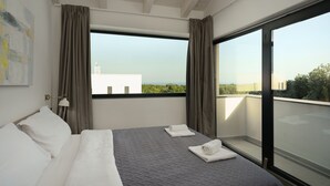 A modern hotel room with a large bed, two pillows, a grey quilted bedspread, and two rolled towels. There is a large window with a view of greenery and buildings. - Villa Krnica (Marcana)