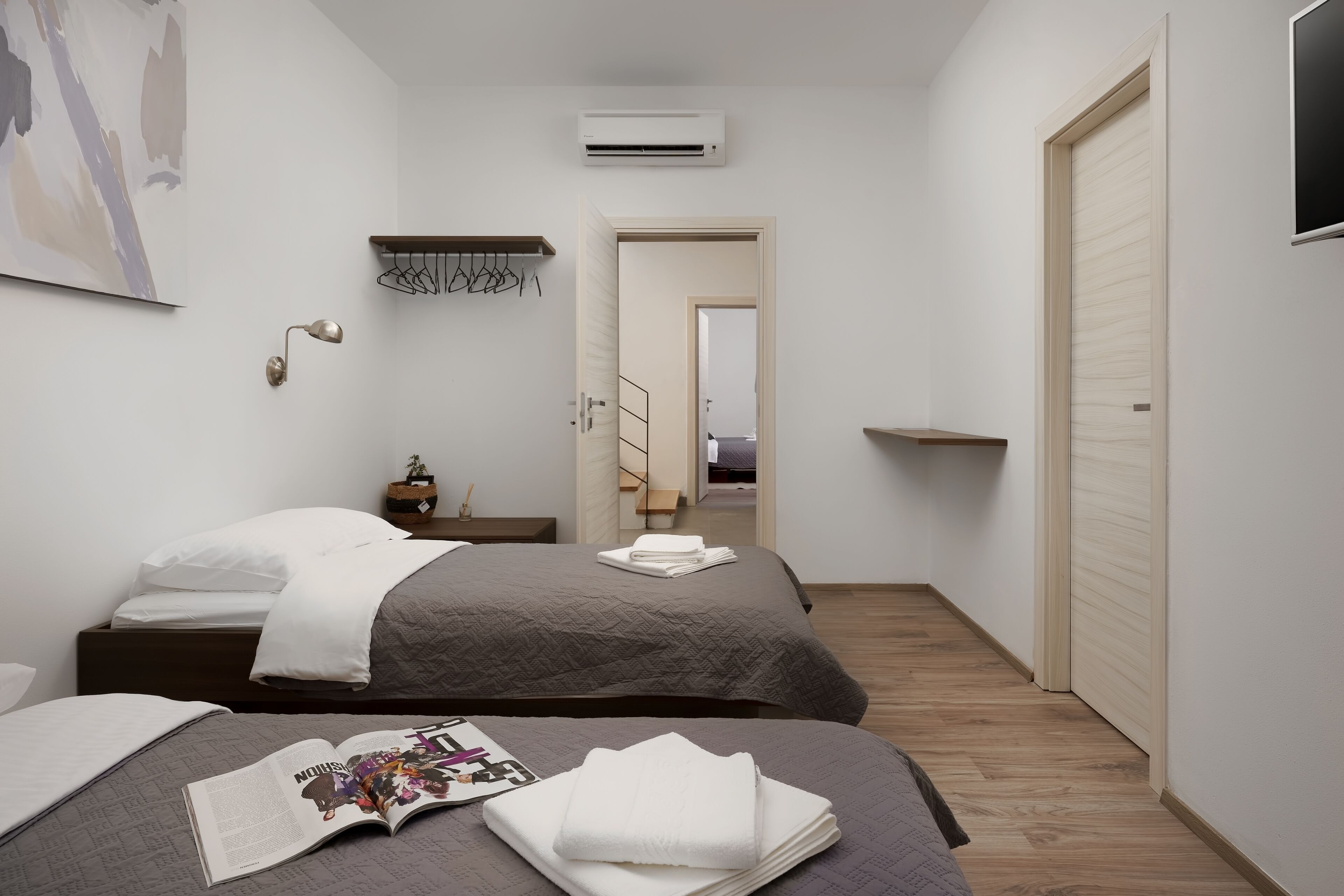 Un dormitorio moderno con dos camas, una mesita de noche, una lámpara de pared y una puerta que da a otra habitación.