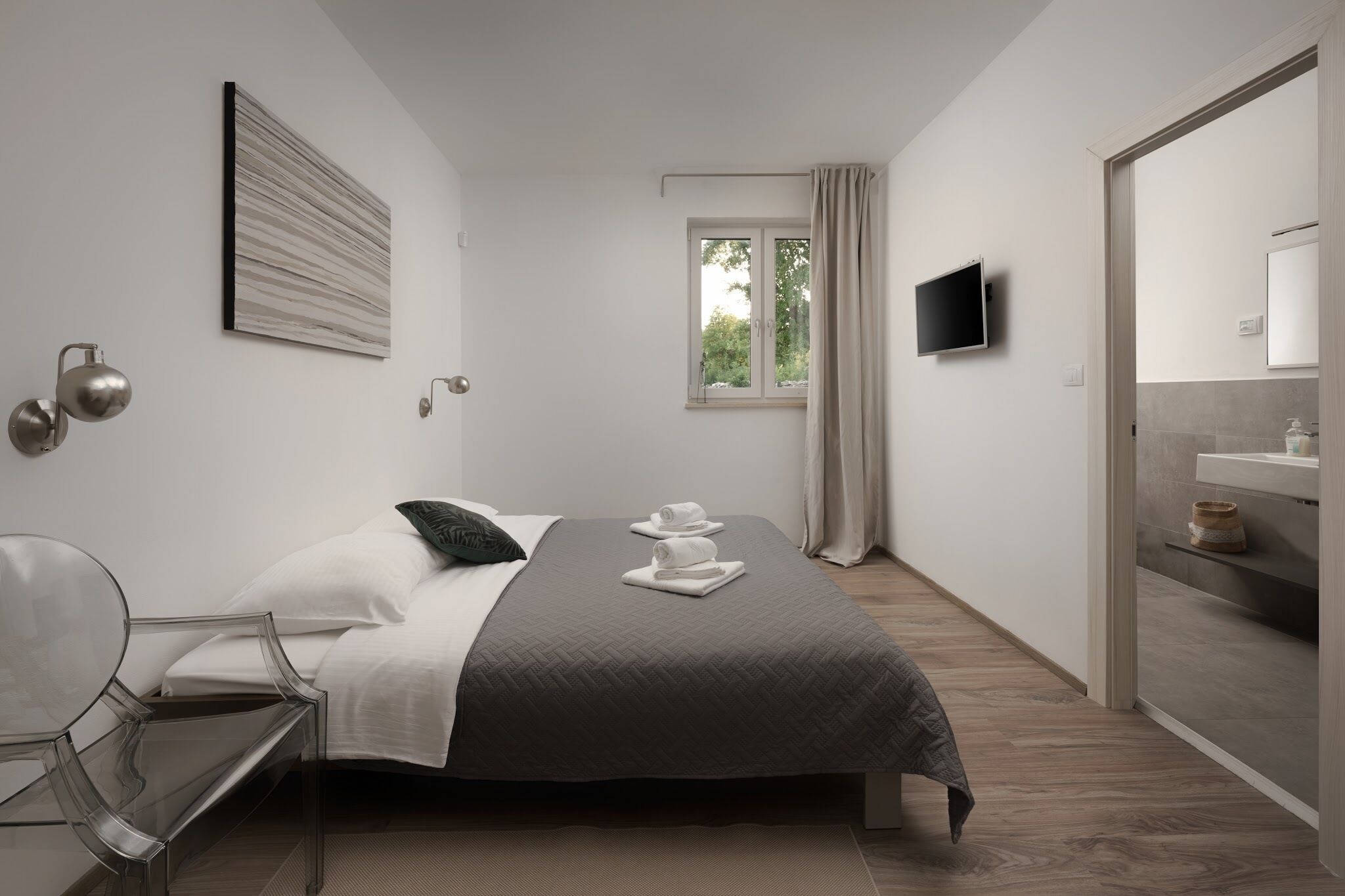 A modern hotel room with a bed, a TV, a window with curtains, a bedside table, and a mirror.