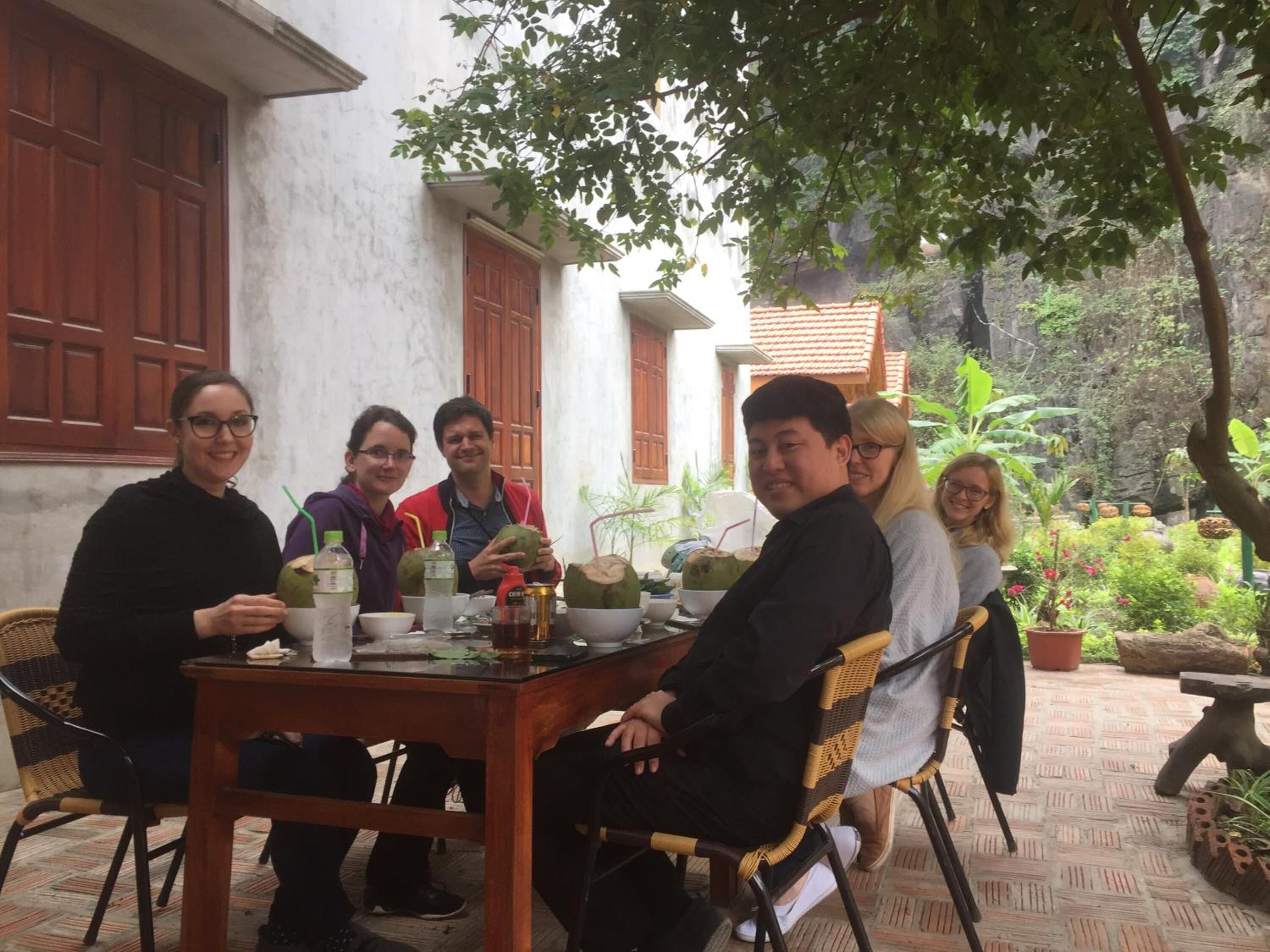 A group of people seated around a wooden table with drinks and food, in an outdoor setting with plants and a building in the background.