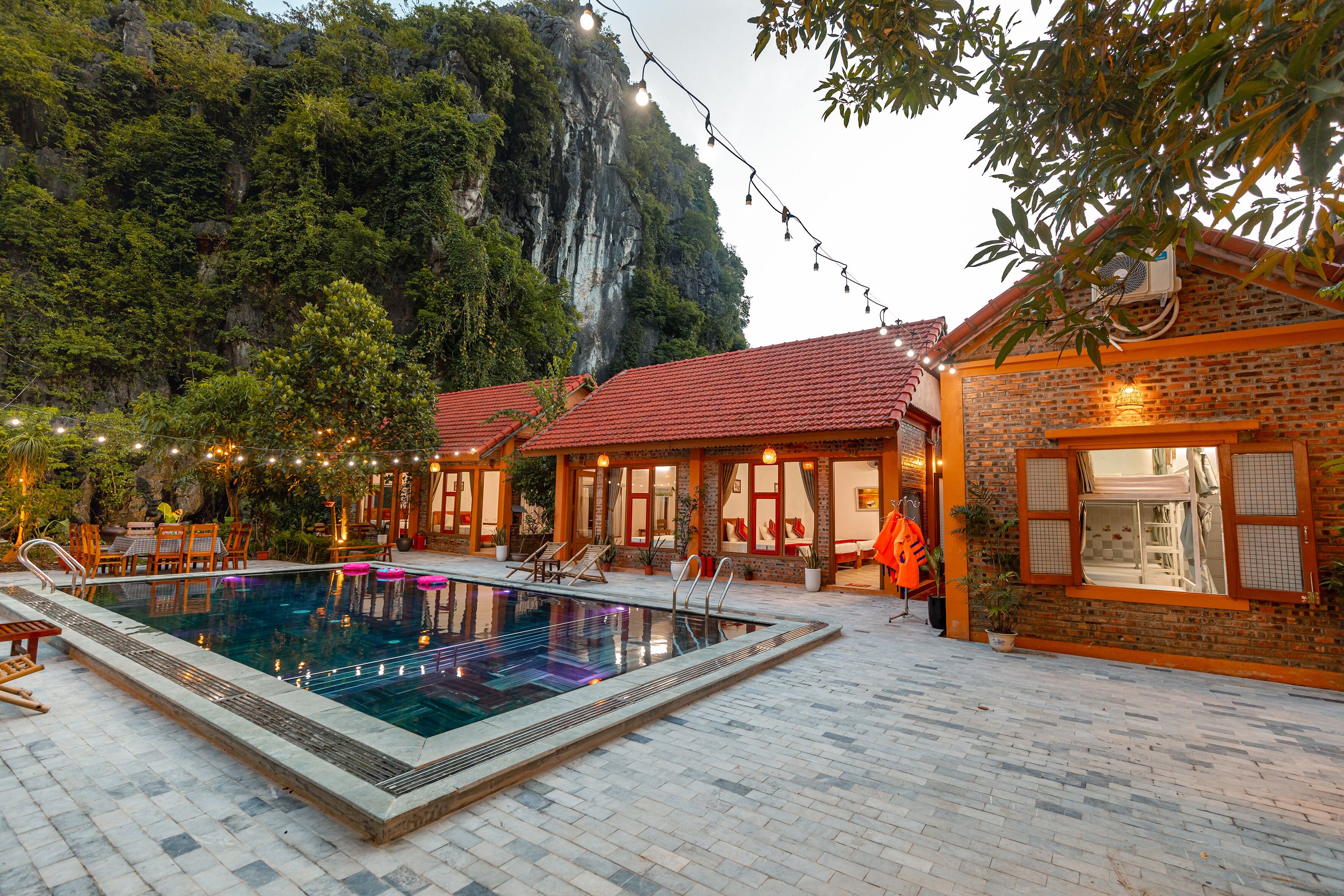 Une piscine avec une terrasse carrelée, entourée d’un bâtiment en briques surmonté d’un toit rouge, et avec une chaîne de montagnes en arrière-plan.