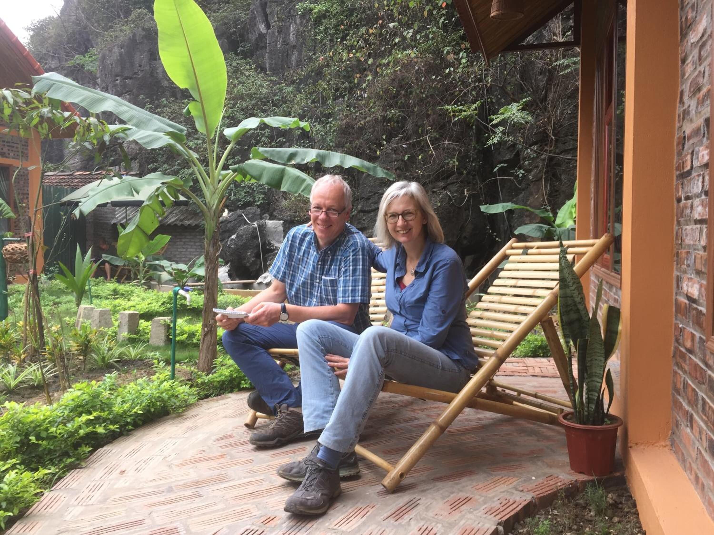 Two people sitting on a wooden swing chair in a garden area with a brick wall and greenery.