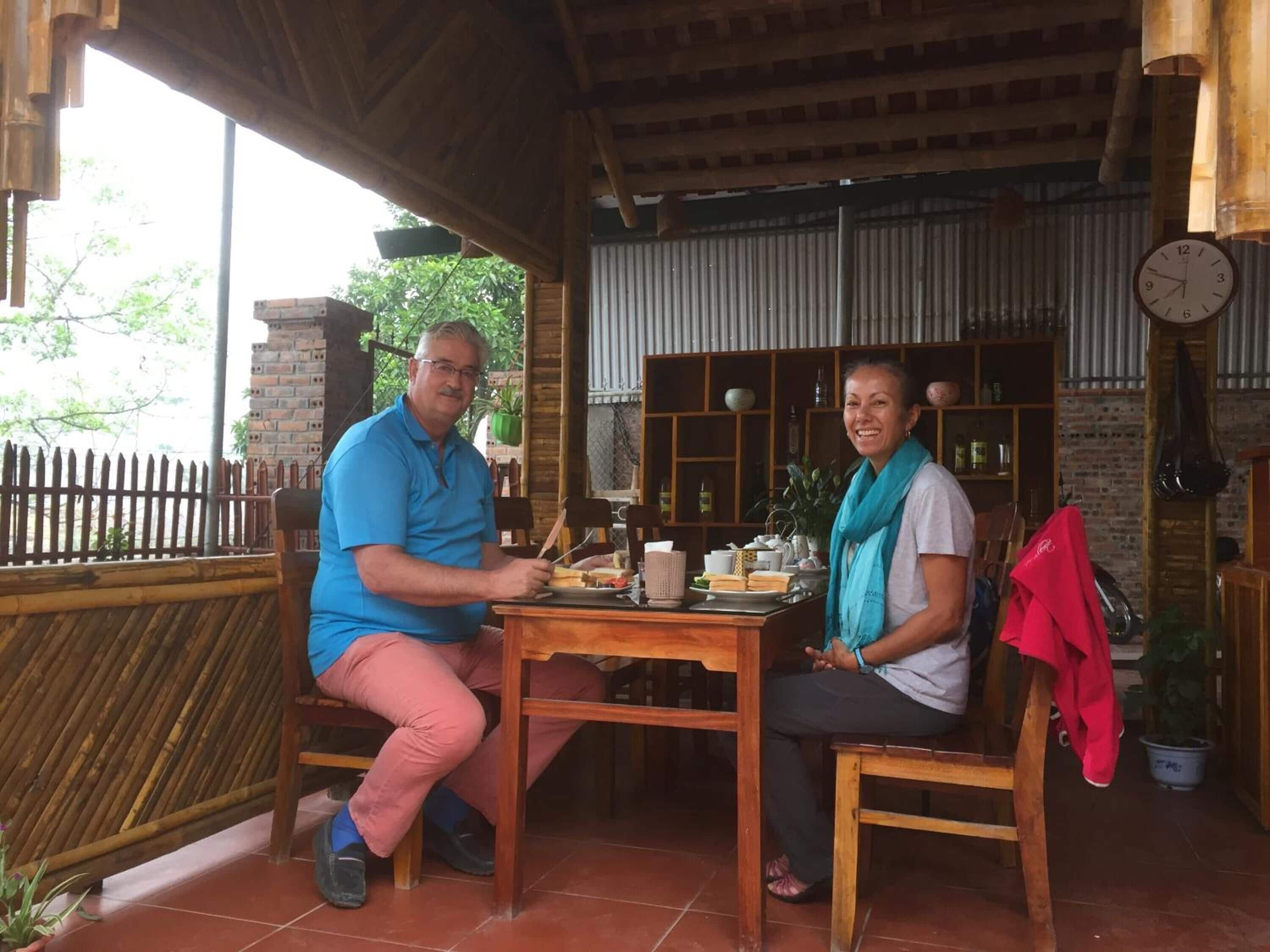 Twee personen zitten aan een houten tafel, achter hen hangt een klok aan de muur.
