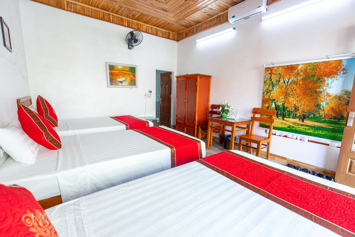 Ein Hotelzimmer mit zwei Betten, Holzbalkendecke, einem Gemälde, einem Ventilator und Blick auf Herbstbäume.