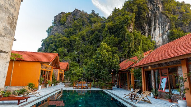 A swimming pool with lounge chairs and potted plants, surrounded by buildings with red-tiled roofs and a mountainous backdrop.