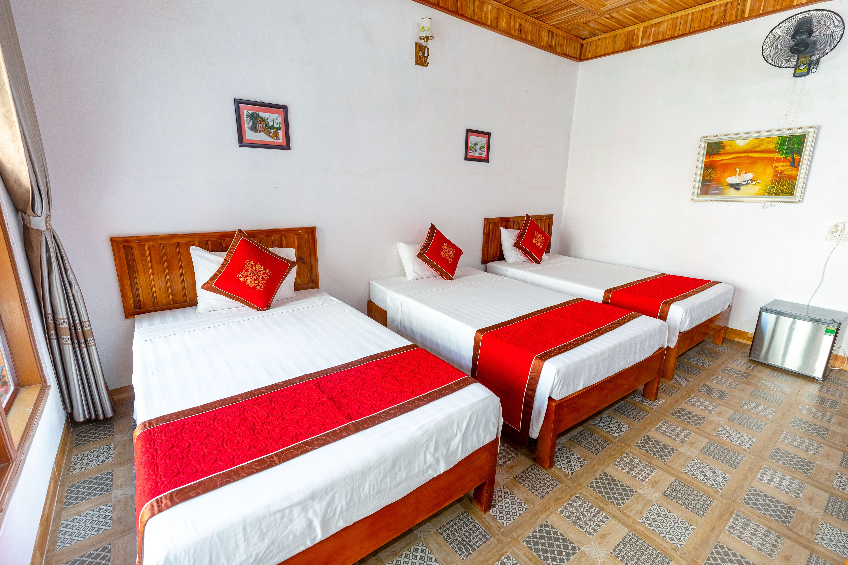 Een hotelkamer met drie eenpersoonsbedden, elk met een rood-wit dekbed en passend kussen. Er is een klein metalen kastje en een ventilator bevestigd aan de muur.