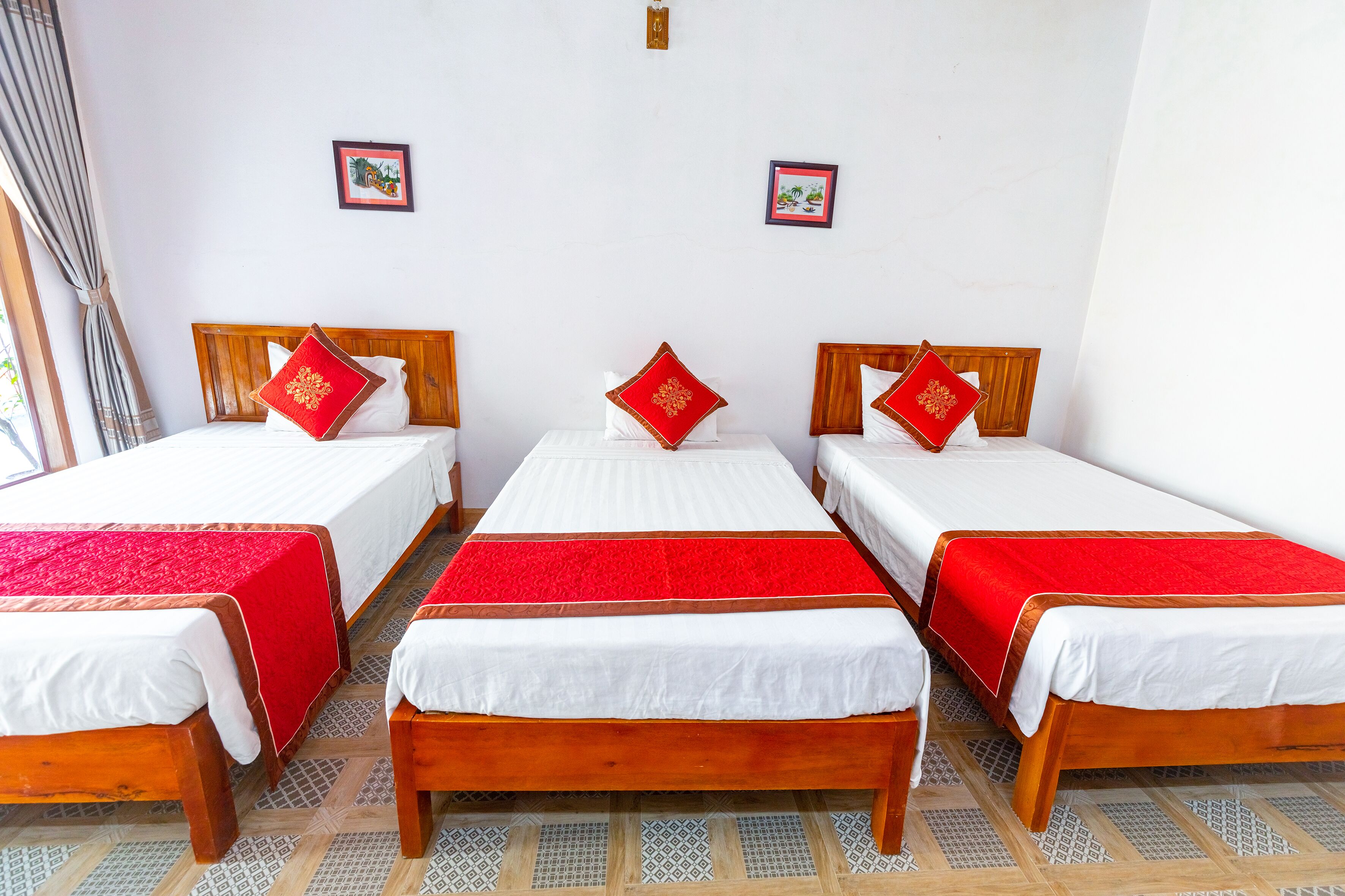 A hotel room with three single beds, each with a wooden headboard and white bedding. The room has a tiled floor and two framed pictures on the wall.