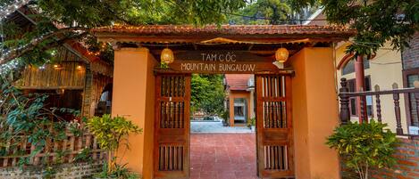 Un arco de madera con un letrero que dice "TAM CÔC MOUNTAIN BUNGALOW" que da acceso a un patio con plantas en macetas y suelo de baldosas.