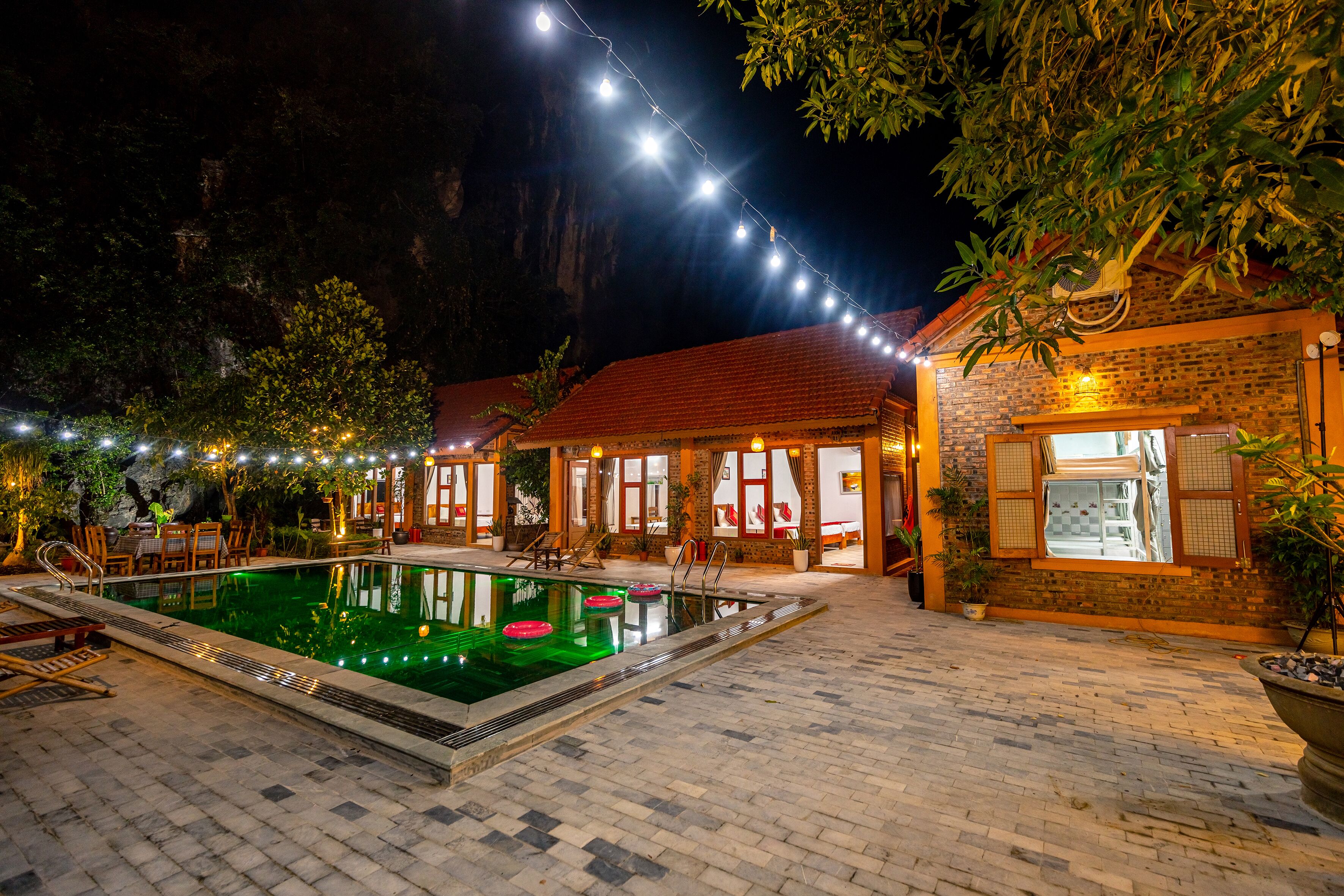 Un área de piscina al aire libre con un patio de ladrillo, sillas de descanso y luces colgantes.