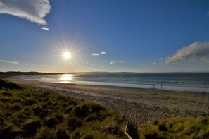 Une plage où le soleil se couche à l’horizon, avec des dunes herbeuses et une mer calme.