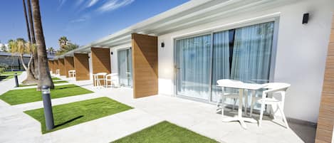 A modern hotel with a patio area featuring white tables and chairs, a well-maintained lawn, and a clear blue sky.