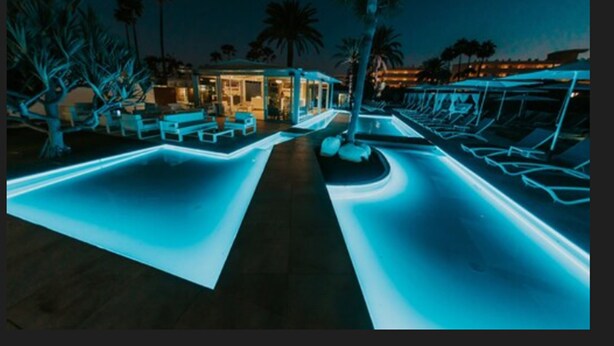 A pool area with illuminated edges, lounge chairs, and a building with glass doors.