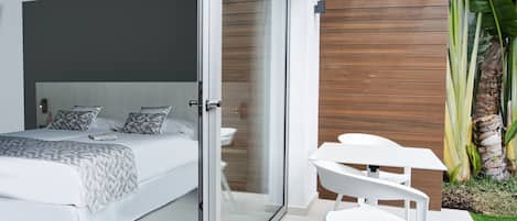 Une chambre moderne dotée d’un grand lit, d’une porte vitrée donnant sur une salle de bain et d’un petit espace extérieur aménagé avec une table et des chaises.