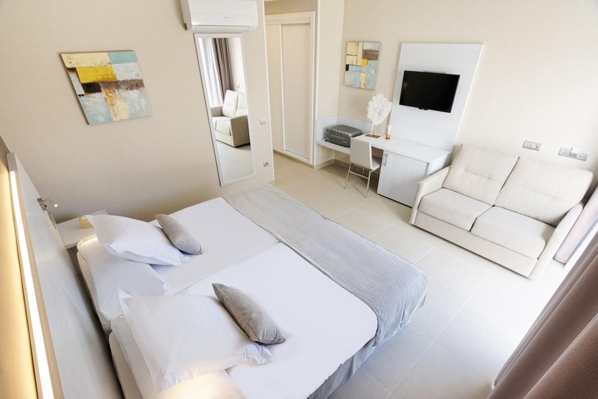 A hotel room with two beds, a sofa, a desk, a TV, and artwork on the walls.
