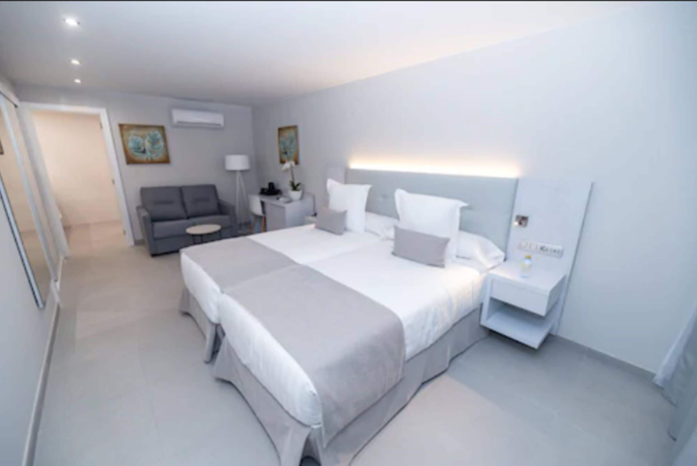 A modern bedroom with a large bed, bedside tables, a grey sofa, and a small table with a lamp.