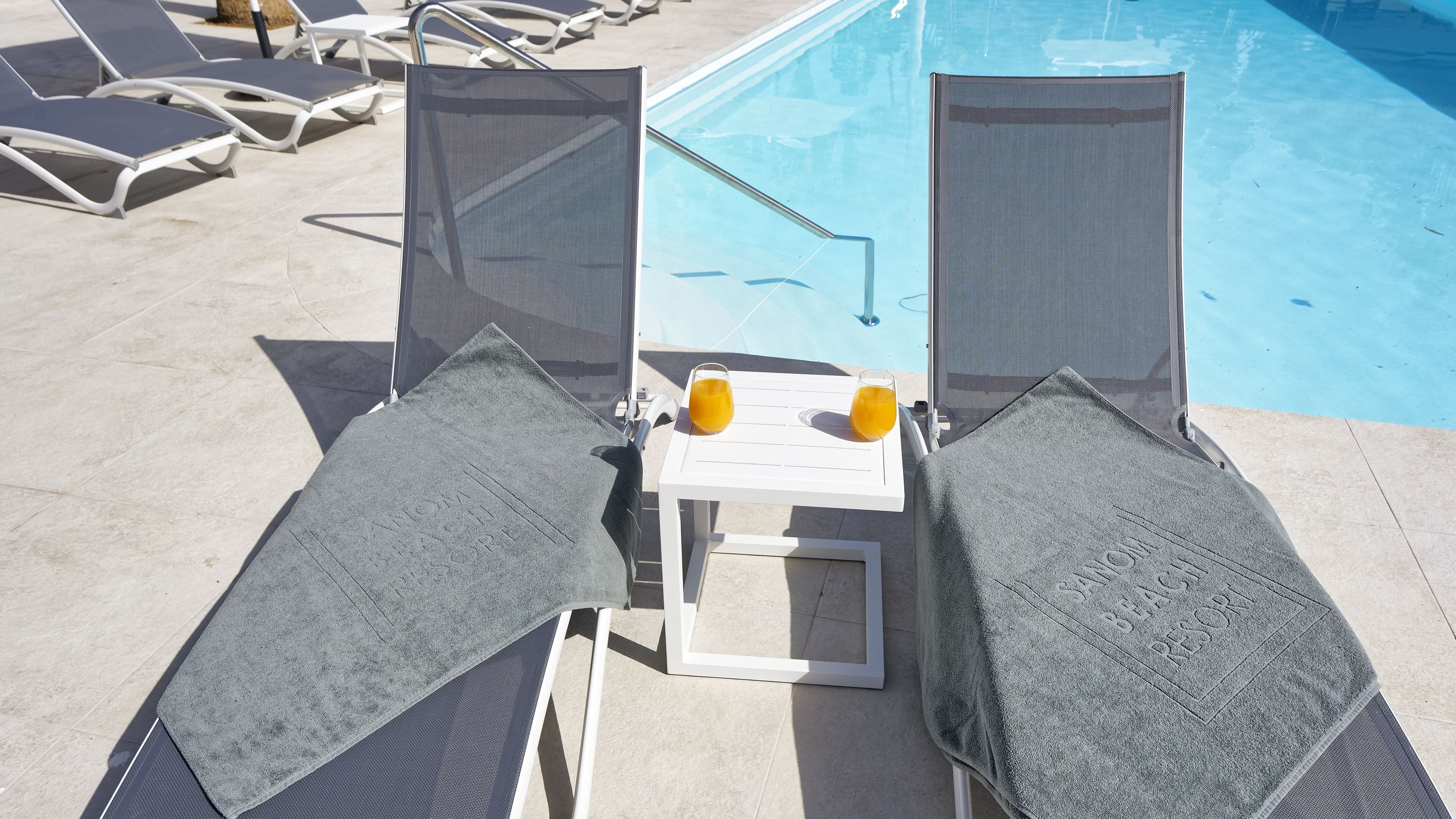 Un área junto a la piscina con dos tumbonas, una mesa pequeña con dos vasos de jugo de naranja y una piscina de agua cristalina.