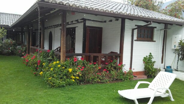 A white building with a tiled roof, a porch with wooden railings, and a garden with colorful flowers. - The Bridge To Ecuador (Pomasqui)
