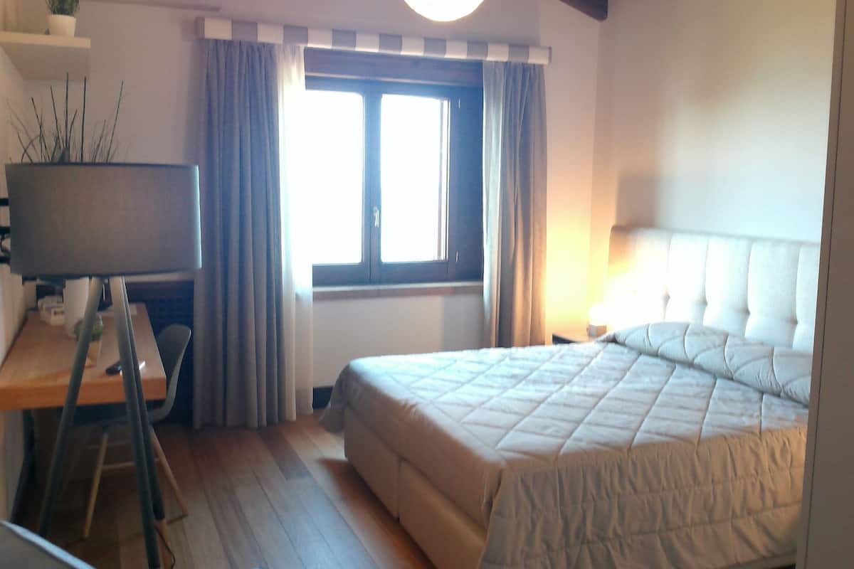 A bedroom with a bed, a desk, a chair, a lamp, and a window with curtains.