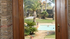 A wooden door with a glass panel, leading to a pool area with palm trees and a clear sky. - CasaRealia (Agrigento)