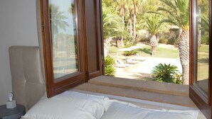 A bedroom with a large bed, a nightstand, and a view of a garden with palm trees. - CasaRealia (Agrigento)