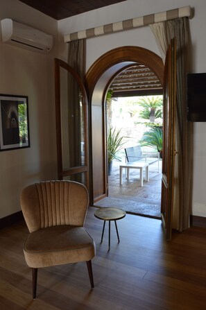 Quarto com piso de madeira, uma poltrona bege, uma mesa redonda pequena e um espelho grande. Há uma porta que dá para uma área externa com um banco e plantas.