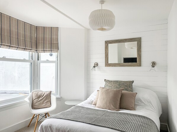 A bedroom with a bed, a chair, a window with blinds, a hanging light fixture, and a mirror on the wall.